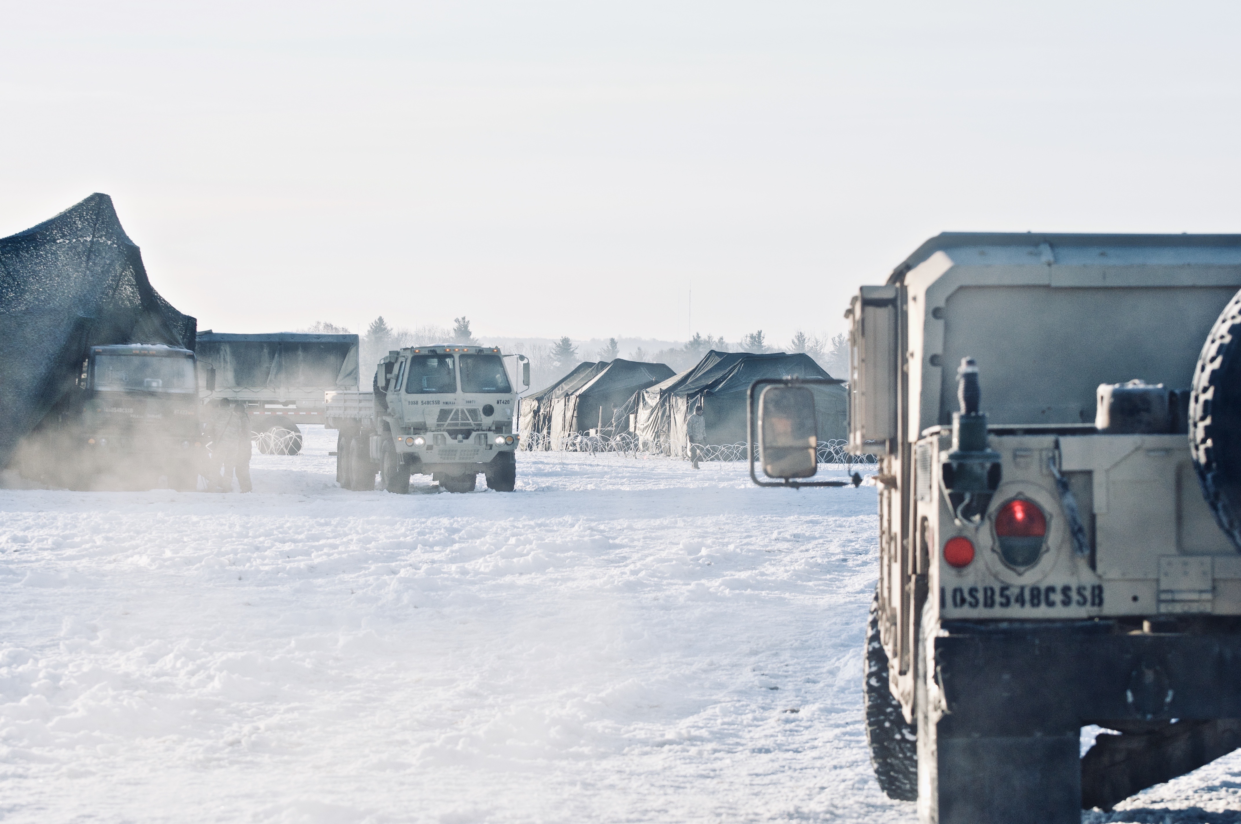 U.S. Army 10th Mountain Division winter encampment at sunrise — convoy trucks and Humvees marked 10SB 548 CSSB amid camouflaged tents in deep snow