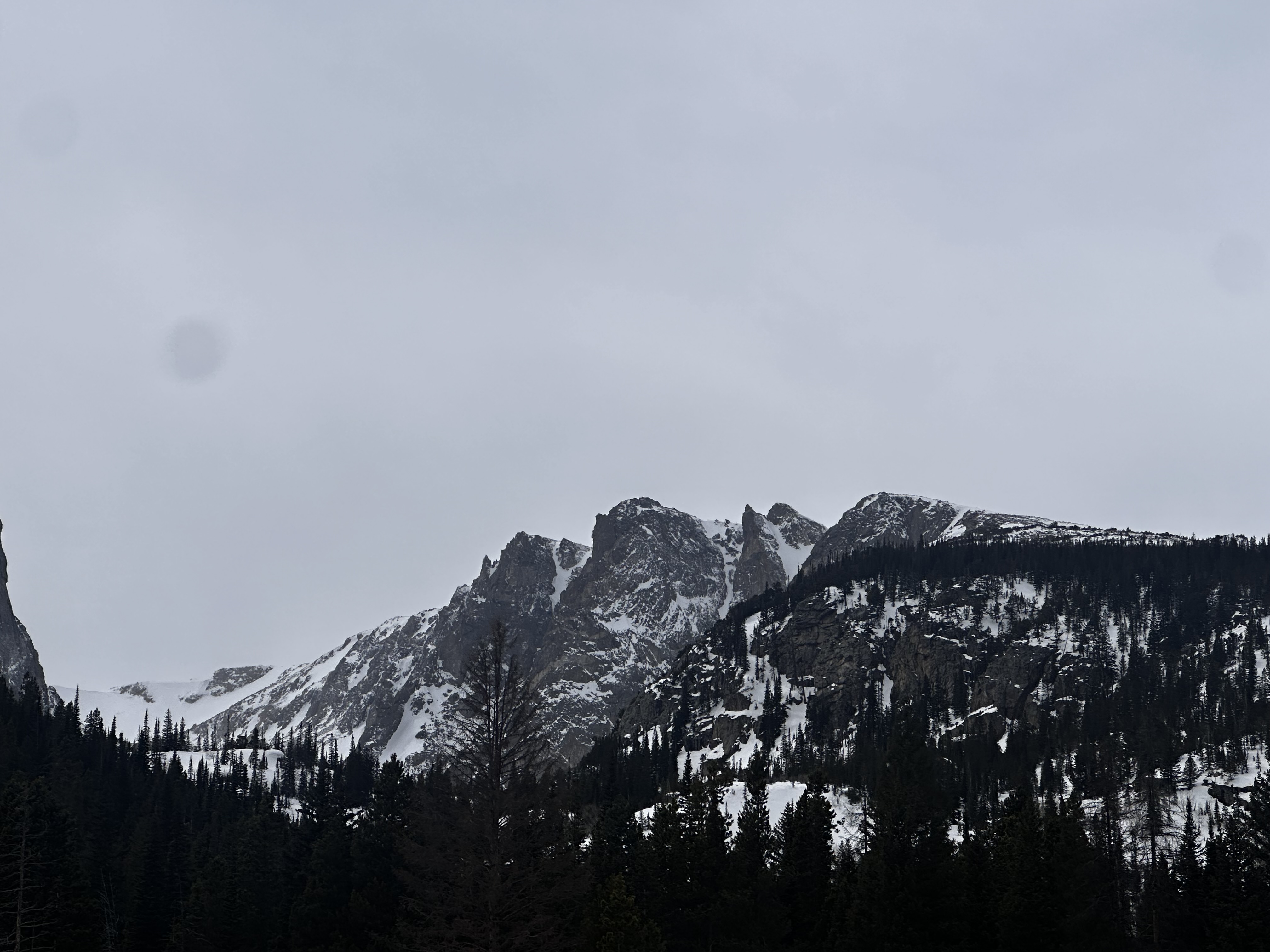 Colorado Front Range peaks under a heavy gray sky, snow-streaked rock and dark evergreen below — the arrival at Restoring the Soul