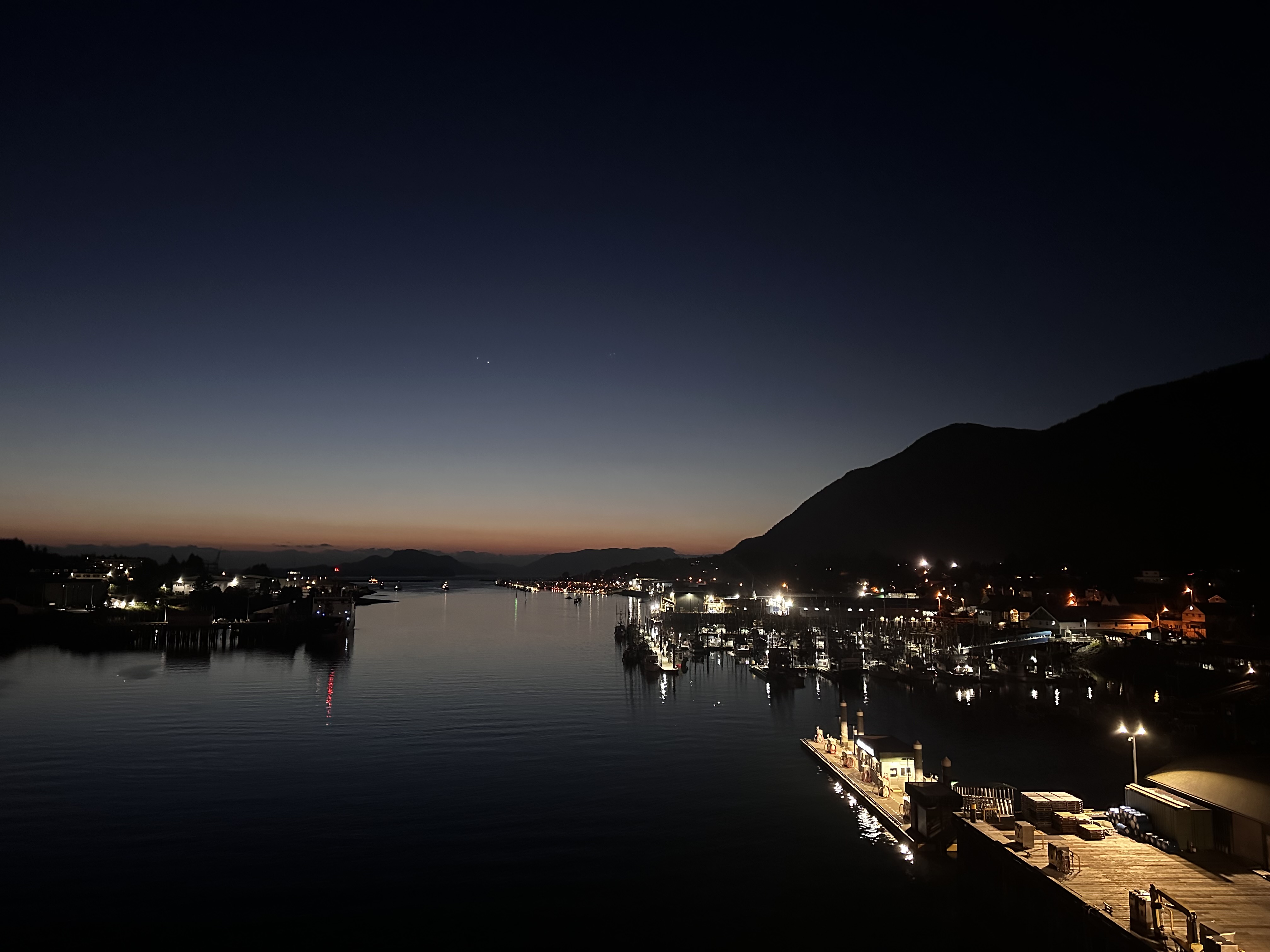 Sitka harbor at twilight from above — dark sky transitioning to a thin warm horizon, working fleet lit at the docks, mountain silhouette to the right